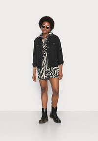 Jaqueta de jeans preta sobre um vestido estampado com um design abstrato em creme e verde, combinado com botas pretas de atacador e óculos de sol.