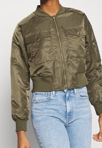 Veste bomber vert olive avec fermeture éclair à l'avant, deux poches poitrine, poignets et ceinture côtelés ; fabriquée dans un matériau lisse et léger.
