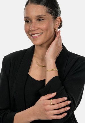 Gold jewelry featuring a chain necklace, bracelet, and ring. Smooth texture and shiny finish, displayed with a black blazer and top.