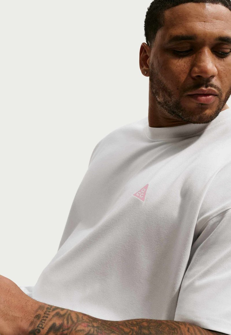 Homme aux cheveux courts et barbe, portant un T-shirt blanc avec un petit logo triangle rose sur la poitrine, regardant vers le bas sur sa gauche.