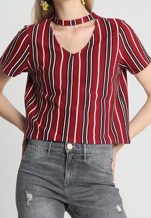 Vrouw draagt een rood, zwart en wit verticaal gestreept V-hals topje met chokerkraag en grijze versleten spijkerbroek, handen rustend op de heupen.