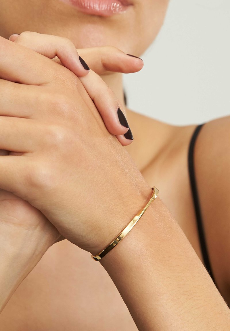 Braccialetto d'oro con una superficie liscia e lucida, indossato su un polso. Presenta un design snodato e arrotondato con una leggera texture martellata.
