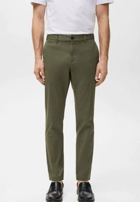 Mężczyzna ubrany w oliwkowozielone, dopasowane spodnie, białą koszulkę i czarne skórzane mokasyny, stojący na tle jednolitego jasnego tła.