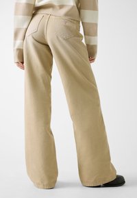 Beżowe spodnie o szerokich nogawkach wykonane z miękkiej bawełny. Posiadają tylne kieszenie oraz naszywkę z logo nad prawą kieszenią. Gładka faktura z delikatnym przeszyciem.