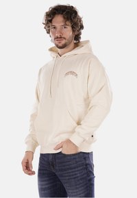 Felpa beige con tasca frontale e cappuccio con cordino, con la scritta "PROPAGANDA" in un design ad arco. Tessuto morbido e vestibilità rilassata.