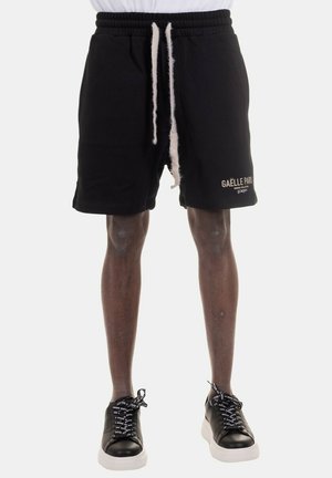 Shorts neri in cotone con vita elastica e stringa, dotati di un logo bianco e testo sulla gamba sinistra; abbinati a sneakers nere.
