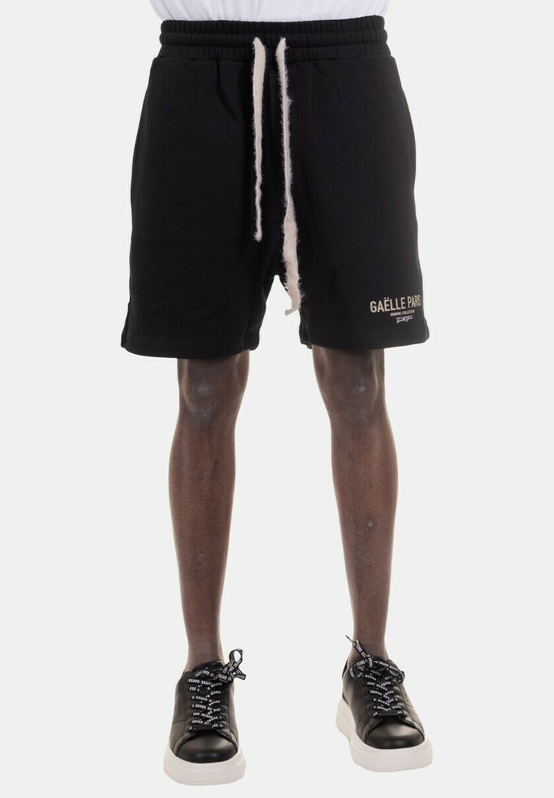 Shorts neri in cotone con vita elastica e stringa, dotati di un logo bianco e testo sulla gamba sinistra; abbinati a sneakers nere.
