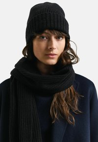 Beanie e sciarpa a coste nere, indossati con un cappotto blu navy. Il cappello è aderente e la sciarpa è spessa e avvolta attorno al collo.
