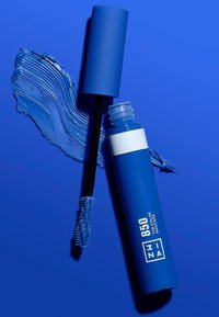 Blå maskara tube med matte finish, hvit etikett og svart børste. Akselert av et strøk med blå maskara. Alle elementene satt mot en blå bakgrunn.