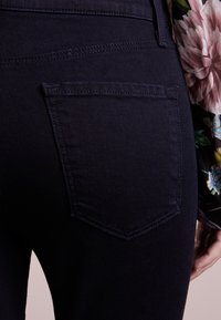Jeans de mezclilla azul oscuro que presentan un bolsillo trasero con un contorno cosido, textura suave y un ligero brillo. Tela floral parcialmente visible.