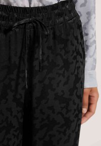 Zwarte sweatpants met een gestructureerd camouflagemotief, een elastische tailleband en een contrasterende trekkoord. De stof lijkt zacht en licht glanzend.