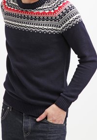 Marinblå tröja med röd, vit och svart nordisk mönstring över bröstet och axlarna, kombinerad med mörka jeans och handen i fickan.