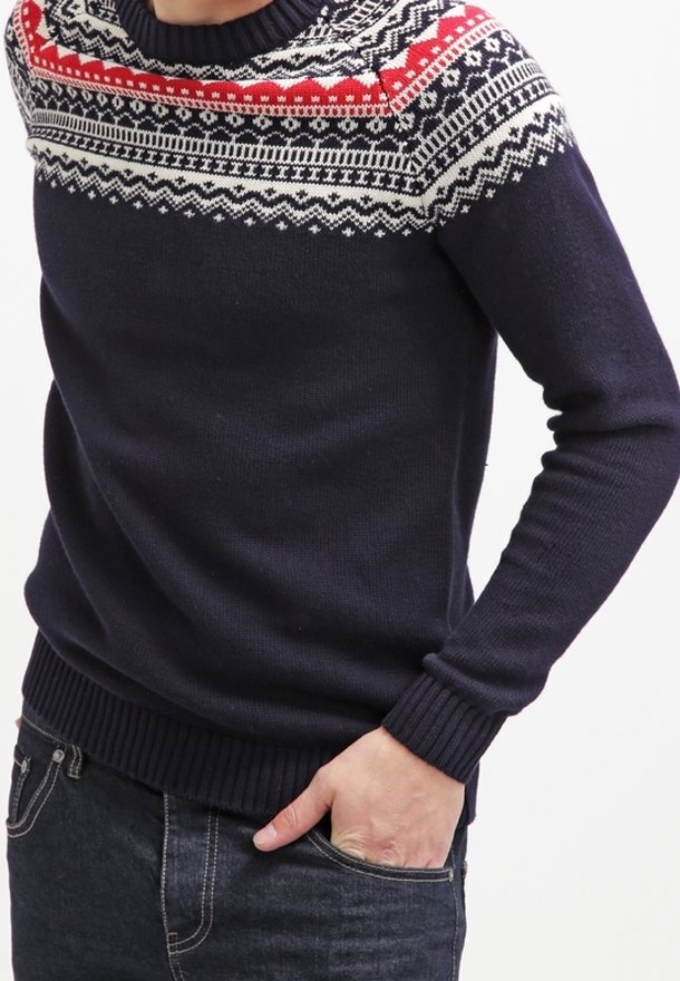 Marinblå tröja med röd, vit och svart nordisk mönstring över bröstet och axlarna, kombinerad med mörka jeans och handen i fickan.