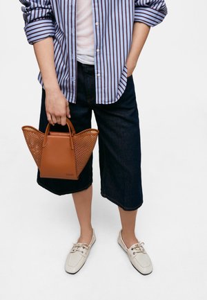 Persona con camisa de rayas remangada, pantalones cortos negros hasta la rodilla, mocasines blancos, sosteniendo un bolso marrón tejido por un asa.