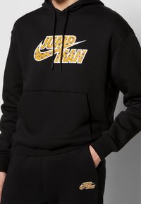 Sort hættetrøje med snøre, der har et stort gult og sort mønstret logo. Kangurulomme og ribstrikkede manchetter fuldender designet.