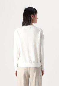 Maglione bianco a maniche lunghe con colletto e polsini a costine, caratterizzato da una texture liscia e da un design aderente, abbinato a pantaloni beige.