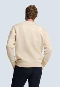 Beige sweatshirt met ribgebreide mouwen en zoom, met witte geprinte tekst en grafiek op de achterkant. Zachte stof met een casual pasvorm.