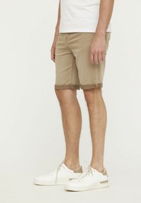 Beige Denim-Shorts mit umgeschlagenen Säumen, ausgestattet mit Vorder- und Gesäßtaschen, kombiniert mit weißen Sneakers mit hellbeigen Akzenten.