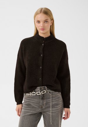 Kvinde med glat blondt hår iført sort knap-op cardigan og grå jeans med et sølv kædebælte mod en ensfarvet baggrund.