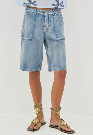 Svetlomodré denimové šortky po kolená s veľkými prednými vreckami, nosené s béžovými sandálmi na členky s cvočkami na nohách osoby.