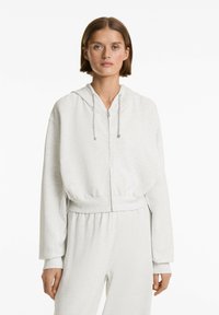 Svetlošedá cropped mikina vyrobená z mäkkého materiálu. Obsahuje zips vpredu, nastaviteľnú kapucňu so šnúrkou a rebrované manžety s uvoľneným strihom.