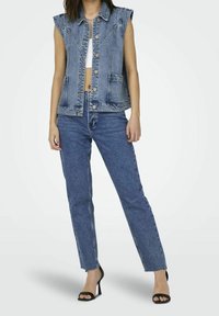 Džínová vesta s krátkými rukávy, zapínáním na knoflíky a dvěma předními kapsami, doplněná o džíny s vysokým pasem v modré barvě a černé podpatky.