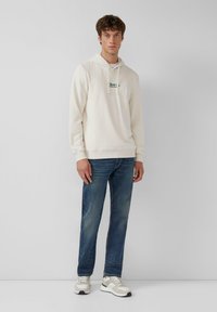 Joven de pie que lleva una sudadera crema con logo, jeans azules y zapatillas blancas frente a un fondo gris liso.