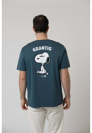 Mann trägt ein türkisfarbenes T-Shirt mit einem mürrischen Snoopy-Cartoon und dem Wort „GRANTIG“ auf dem Rücken, steht vor einfachem Hintergrund.