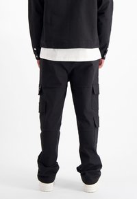 Zwarte cargobroek met meerdere zijzakken, gemaakt van textuurstof. Gedragen met een wit shirt, aangevuld met witte sneakers.