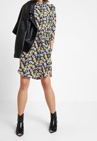 Abito floreale con sfondo nero, caratterizzato da piccoli fiori multicolori, maniche 3/4 e vestibilità ampia, abbinato a una giacca di pelle nera e stivaletti.