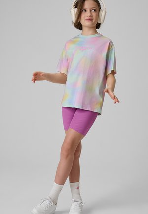 Dziecko w białych słuchawkach, pastelowej koszulce tie-dye z napisem, fioletowych spodenkach rowerowych, białych skarpetkach i białych tenisówkach, stojące w zrelaksowanej pozie.