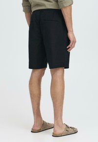 Svart, knelange shorts laget av mykt stoff med elastisk linning og to baklommer, kombinert med beige sandaler.