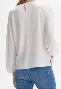 Blouse blanche texturée avec des manches longues et un petit design en trou de clé au dos, assortie à un jean en denim bleu.