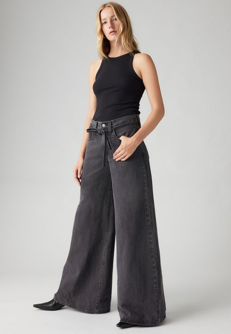 Schwarzes, geripptes Tanktop kombiniert mit weiten, hoch taillierten, dunkelgrauen Jeans, die einen Kordelzug an der Taille und seitliche Taschen haben.