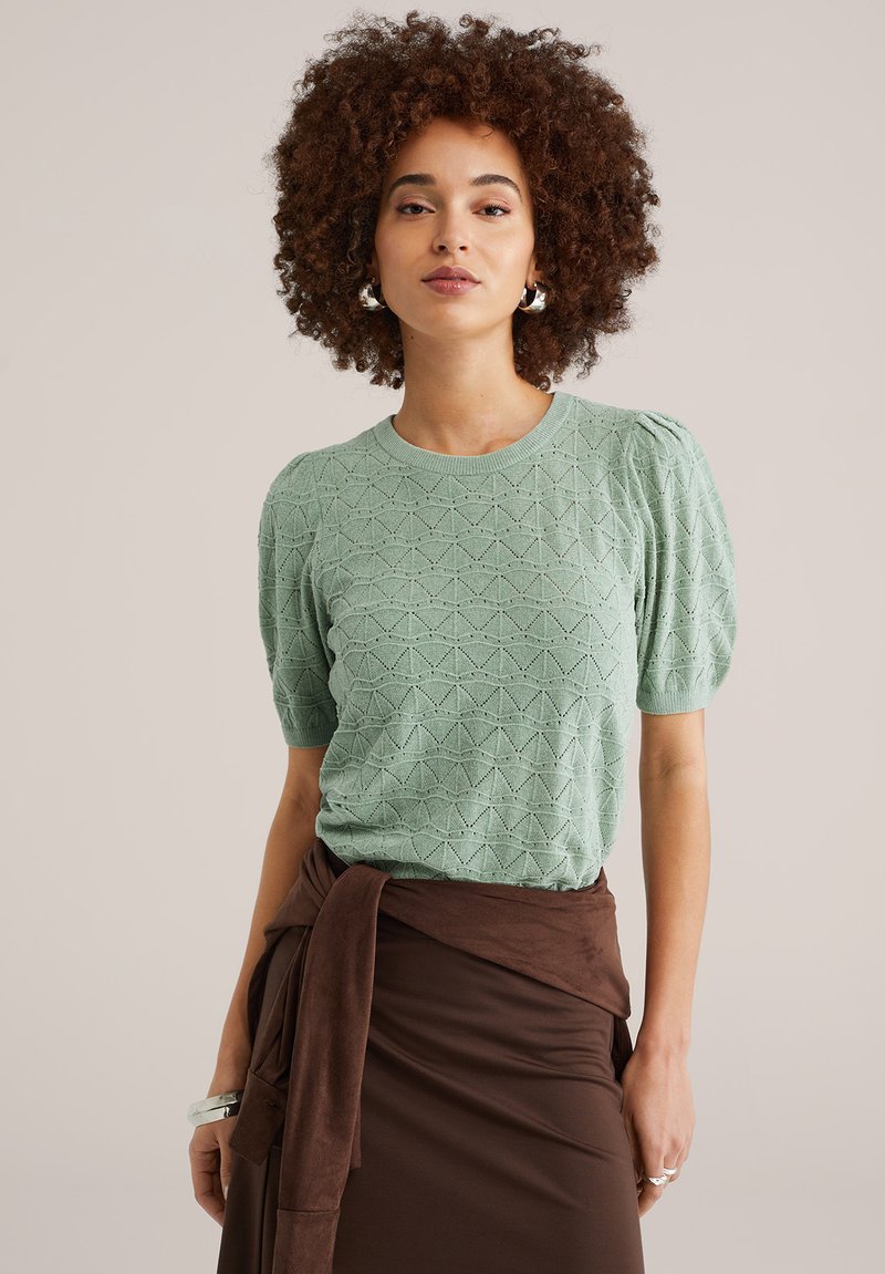 Šviesiai žalias trumpomis rankovėmis megztas megztinis su tekstūriniu geometriniu raštu, derinamas su ruda vyniojama sijonu.