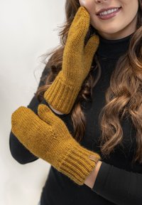 Mosterdgele gebreide wanten met geribde manchetten, textuurstof en een kleine leren labelaccent. Draagt door een persoon tegen een neutrale achtergrond.