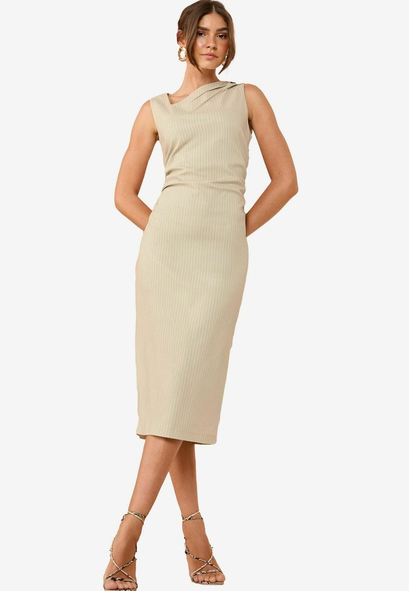Beige ribattu midi dress with a diagonal neckline, fitted shape, ja hienovarainen tekstuuri. Malli on vaaleissa sandaaleissa, kädet taskuissa.