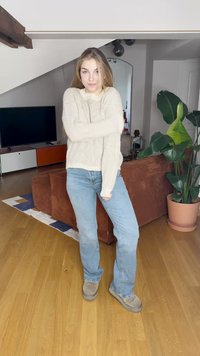 Suéter blanco de punto con textura acanalada, jeans azul claro y botas grises tipo slip-on. Entorno interior con un sofá marrón y una planta verde.