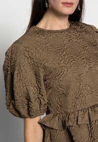 Blusa castanha texturizada com mangas bufantes, apresentando uma bainha com volant e um padrão circular. A costura é visível no tecido.
