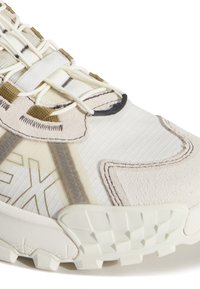 Gros plan d'une basket beige et crème avec une semelle texturée, des matériaux en daim et en tissu, et des lacets élastiques avec une languette.