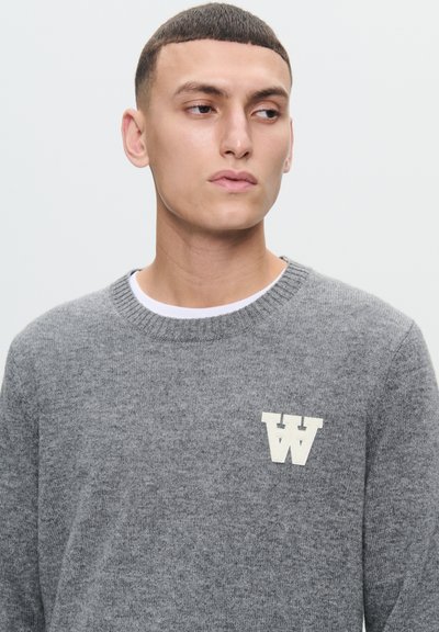 Grå strikket sweater med rund hals, der har et hvidt "W" logo på venstre bryst. Teksturen virker blød og behagelig.