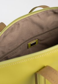 Groene leren tas interieur met een gladde bruine voering, een ritszak en een labeltag. Stevige bruine banden zichtbaar aan de zijkanten.
