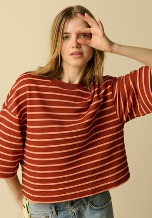 Jeune femme aux cheveux blonds portant une chemise rayée rouge et rose, formant un cercle avec ses doigts autour de son œil droit.