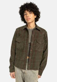 Ragazzo con capelli ricci che indossa una camicia a quadri verde sbottonata sopra un top corto grigio e jeans chiari su uno sfondo semplice.