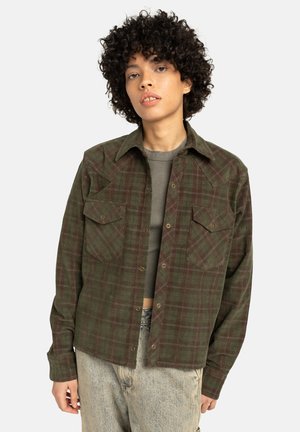 Ragazzo con capelli ricci che indossa una camicia a quadri verde sbottonata sopra un top corto grigio e jeans chiari su uno sfondo semplice.
