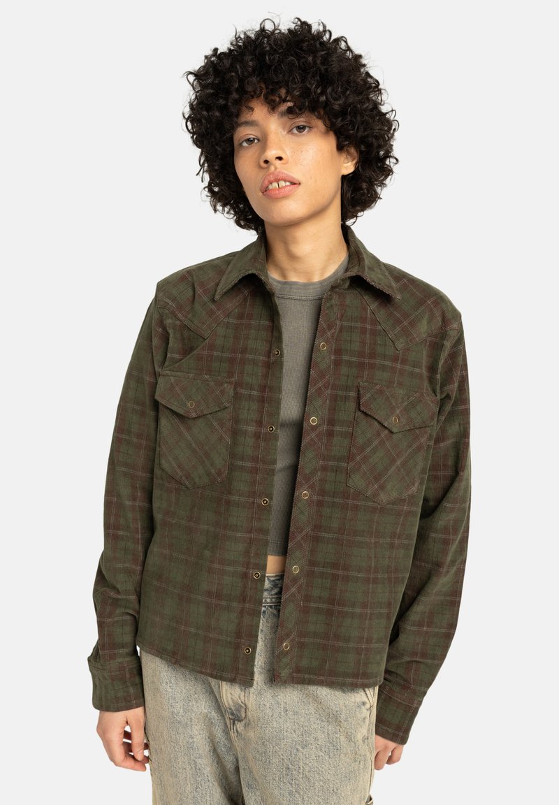 Ragazzo con capelli ricci che indossa una camicia a quadri verde sbottonata sopra un top corto grigio e jeans chiari su uno sfondo semplice.