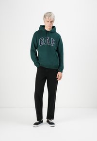 Young person wearing a dark green GAP hoodie, black pants, and black sneakers, standing against a plain white background.