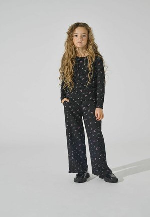Sort ribbet stof sæt med blomsterprint. Langærmet top, vide bukser og sorte platform sko. Model står med den ene hånd i lommen.