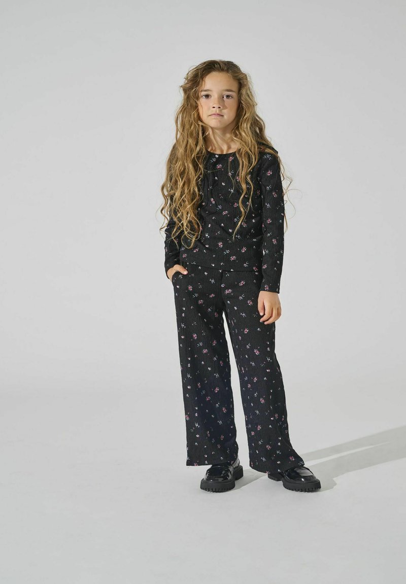 Zwart geribbeld stoffen outfit met bloemenpatroon. Lange mouwen top, wijde broek en zwarte platform schoenen. Model staat met één hand in de zak.