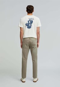 Tricou alb din bumbac cu un imprimeu grafic albastru pe spate, reprezentând o cafetieră, o ceașcă, un croissant și text deasupra. Asortat cu pantaloni verzi deschis, de măsline.